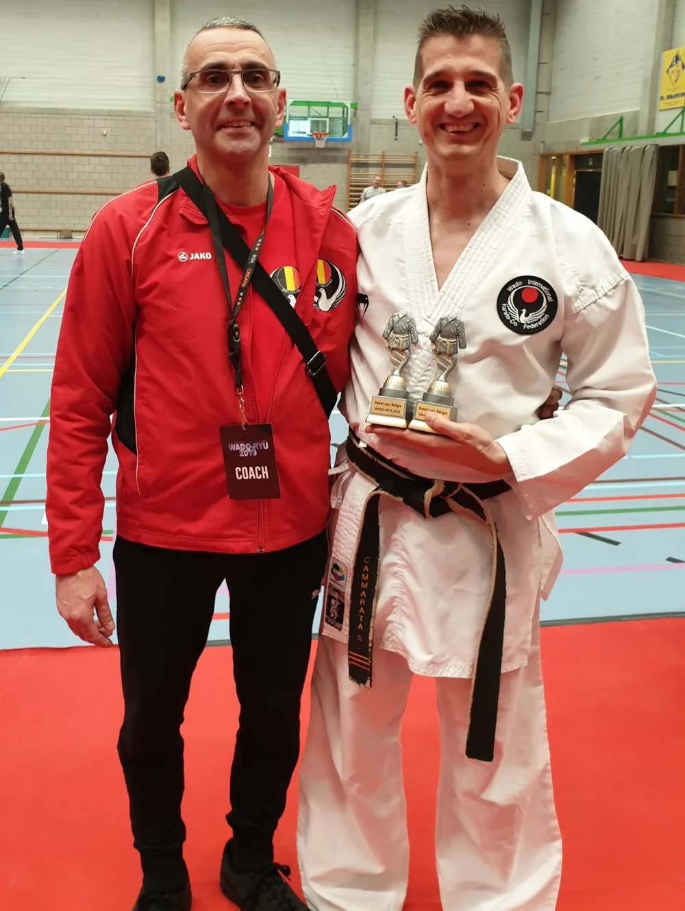 Sandro et Alberto, les deux instructeurs du club, en salle lors d’une compétition : l’un en veste de coach, l’autre en kimono et ceinture noire, deux coupes en main.